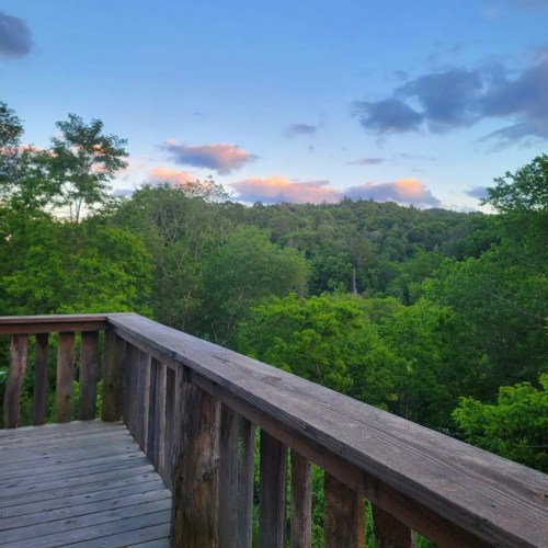 A wooden deck overlooks a lush green landscape with trees and a colorful sky at sunset.