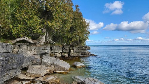 A serene lakeside scene with rocky shorelines, trees, and a clear blue sky with scattered clouds.