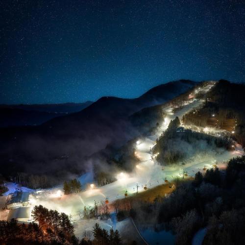 A nighttime aerial view of a ski resort, with illuminated slopes and starry sky above.