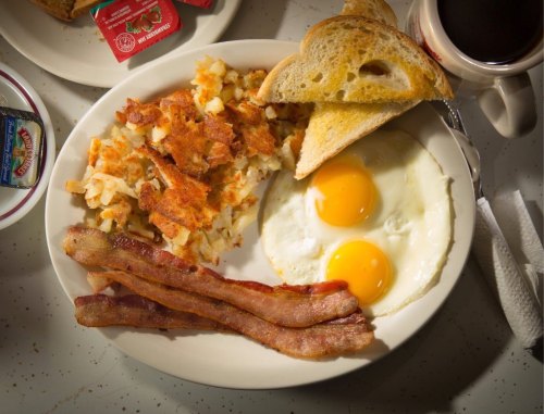 A plate of breakfast featuring two fried eggs, crispy bacon, hash browns, toast, and coffee on the side.