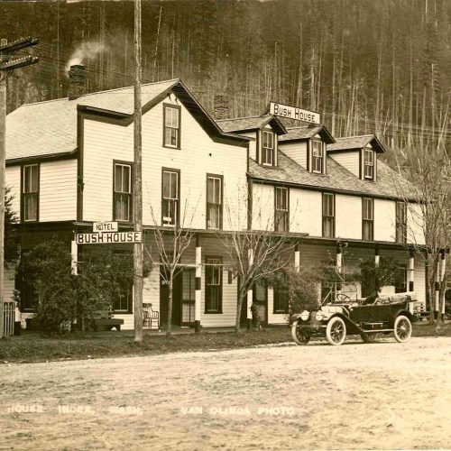 Historic photo of the Bush House hotel with a vintage car parked outside, surrounded by trees and a dirt road.