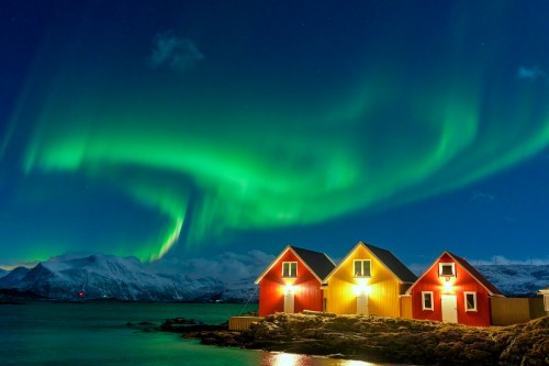 Colorful northern lights illuminate a serene landscape with three cozy cabins by the water, surrounded by mountains.