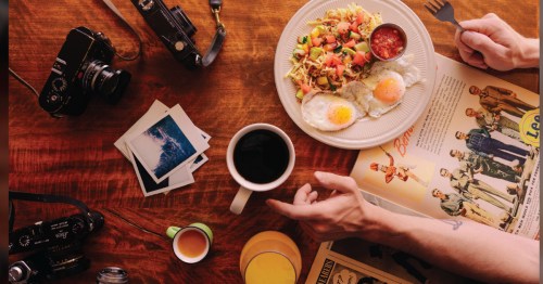 Looking down at a wooden table covered in photos, a camera, coffee, a mimosa, breakfast hash, and a magazine.