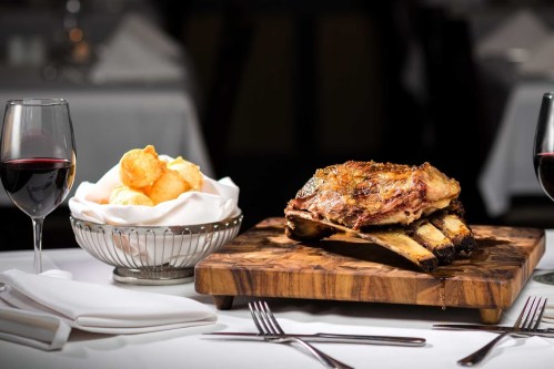 A beautifully plated rack of ribs on a wooden board, accompanied by a basket of bread rolls and two glasses of red wine.