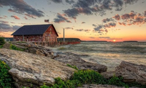 A scenic sunset over a rocky shoreline with a rustic building and gentle waves under a colorful sky.