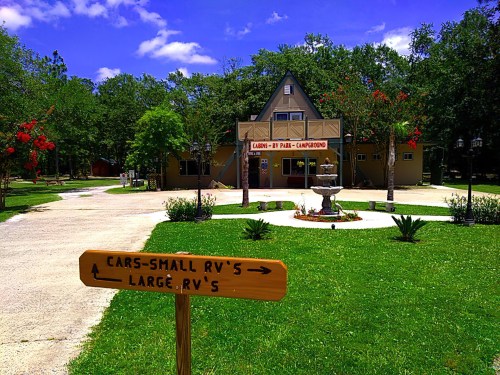 A sign points to parking for cars and RVs in front of a cabin-style building surrounded by greenery and flowers.