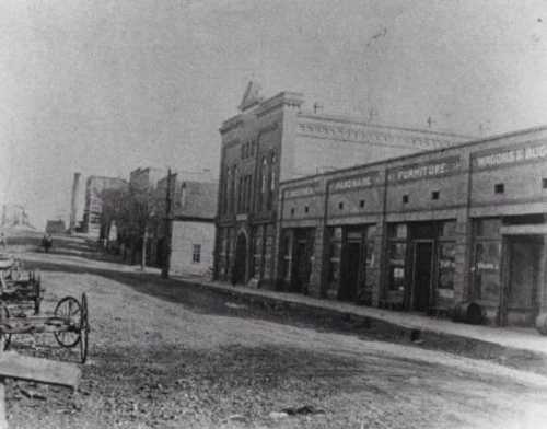 Black and white photo of a dusty street lined with old buildings and wagons, depicting a historic town scene.