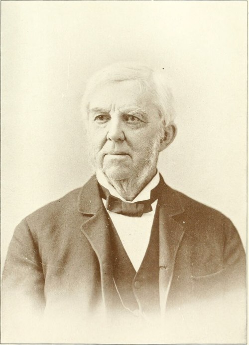 A sepia-toned portrait of an older man with white hair and a beard, wearing a suit and bow tie.