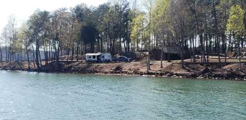 A serene lakeside scene featuring an RV parked near a wooded area with trees and a calm water surface.