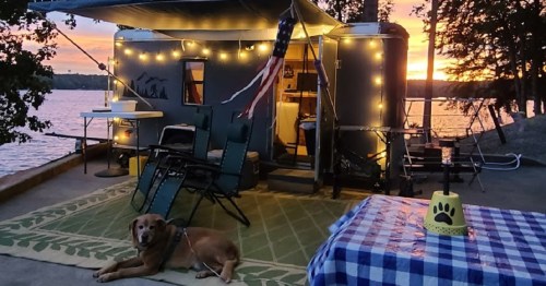 A cozy campsite by the lake at sunset, featuring a trailer, chairs, string lights, and a dog resting on a rug.