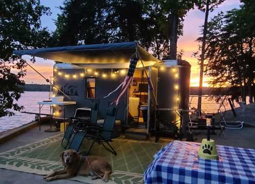 A cozy campsite by the lake at sunset, featuring a trailer, outdoor seating, and a dog resting on a rug.