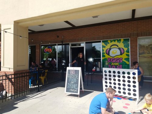 A storefront with an "Open" sign, a chalkboard warning against outside food, and a large Connect Four game outside.
