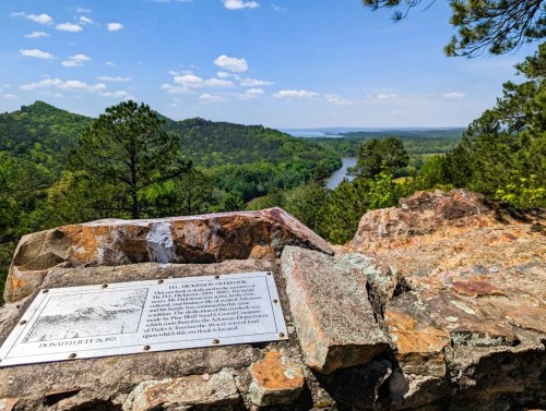 Scenic view from a rocky overlook, featuring a river and lush green hills under a blue sky with clouds.