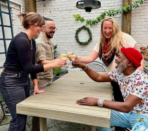 Four friends toast with cocktails at a table in a cozy outdoor setting, smiling and enjoying each other's company.