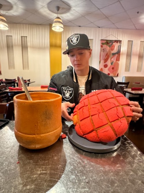 A person sits at a table in a restaurant, preparing a large red concha next to a clay pot.