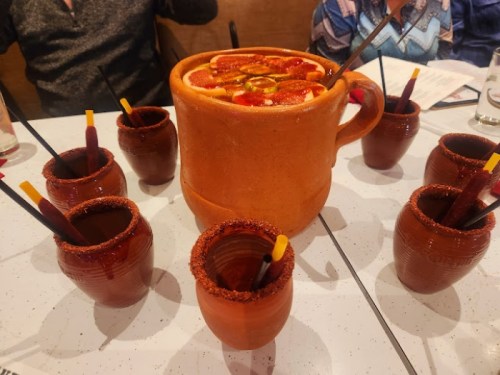 A large clay mug filled with a colorful drink surrounded by smaller clay cups, all with straws and rimmed with chili.