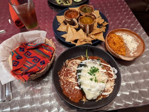 A colorful plate of enchiladas with rice, alongside chips and salsas, with a woven basket of tortillas in the foreground.