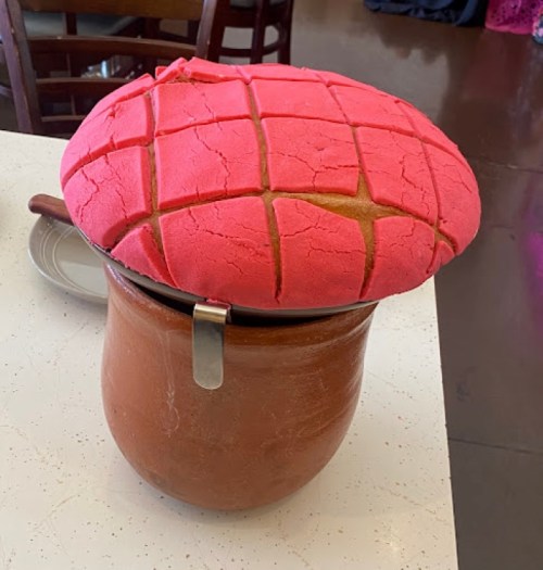 A large, pink concha bread sits atop a clay pot on a table, showcasing its textured, grid-like surface.