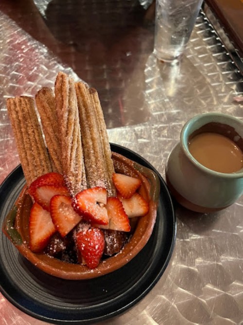A bowl of churros topped with strawberries and powdered sugar, served with a cup of chocolate sauce.