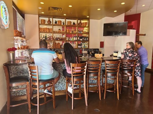 A cozy bar area with patrons seated at the counter, surrounded by shelves of drinks and decorative items.