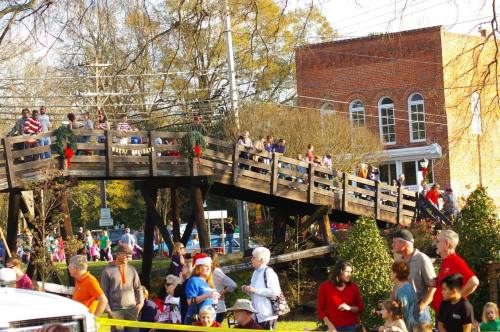 A festive crowd gathers on a wooden bridge, with people in holiday attire and decorations in a small town setting.