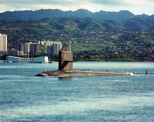 A submarine surfaces in calm waters with mountains and a city skyline in the background.