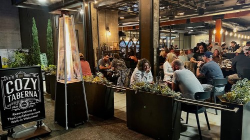 A bustling outdoor dining area with a tall heater, plants, and a sign for "Cozy Taberna" in a lively atmosphere.