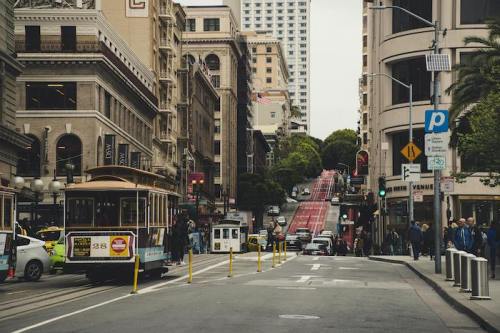 A busy city street with a cable car, traffic, and pedestrians, lined with historic buildings and a steep hill in the background.
