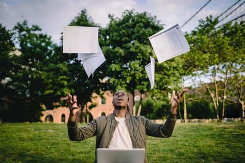 A person sits on grass, joyfully tossing papers into the air with a laptop in front of them and trees in the background.