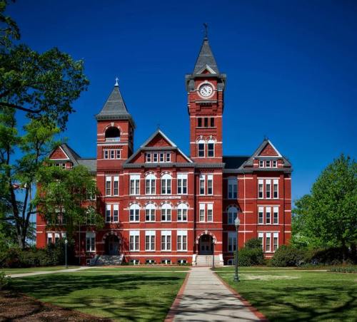 Historic red brick building with a clock tower, surrounded by green trees and a clear blue sky.