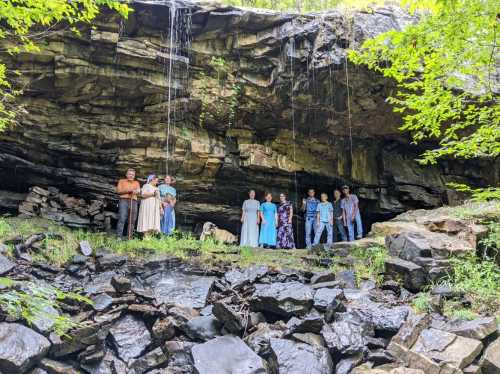 A group of people stands under a rocky overhang with water cascading down, surrounded by greenery and rocks.