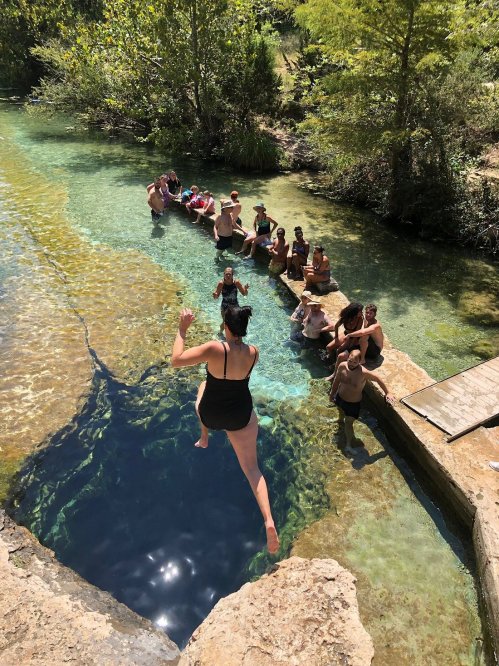 A person jumps into a clear blue pool while others relax on rocks nearby, surrounded by lush greenery.