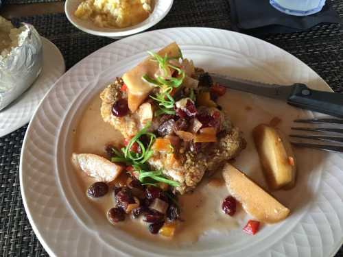 A plate of fried chicken topped with fruit and vegetables, served with baked potato and a side of creamy dish.