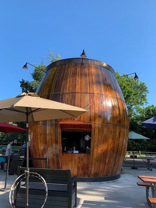 A large wooden barrel-shaped structure with outdoor seating and umbrellas, set against a clear blue sky.