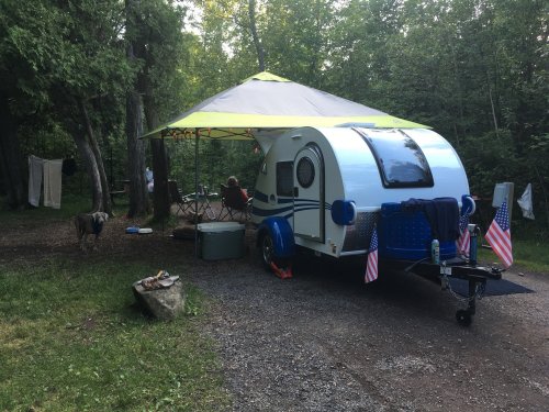 A small blue camper with a canopy set up in a wooded area, featuring a picnic table and a dog nearby.
