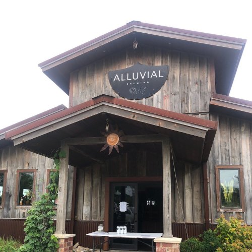 A rustic wooden building with a sign reading "Alluvial" and a welcoming entrance surrounded by greenery.