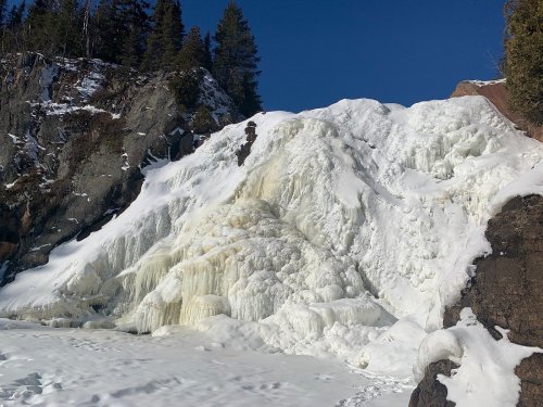 Frozen waterfall cascading down rocky cliffs, surrounded by snow and evergreen trees under a clear blue sky.