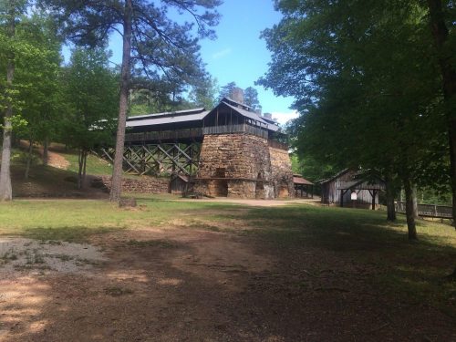 A rustic building with a stone base and wooden upper structure, surrounded by trees and a grassy area.
