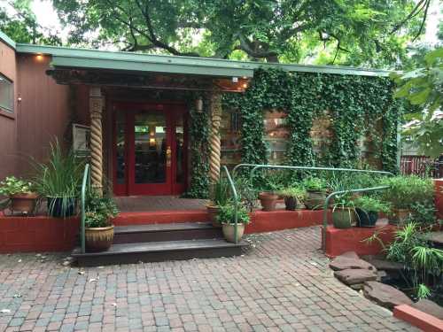 A charming restaurant entrance surrounded by greenery and potted plants, featuring red doors and stone steps.