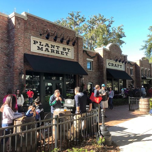 A bustling outdoor market scene with people gathered in front of brick buildings labeled "Plant St. Market" and "Craft."