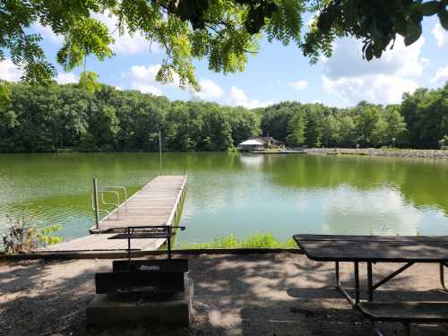 A serene lake view with a wooden dock, surrounded by lush greenery and a picnic area in the foreground.