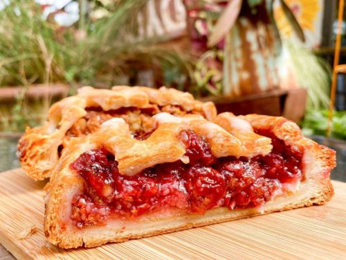 A close-up of a sliced cherry pie on a wooden board, showcasing its flaky crust and vibrant red filling.