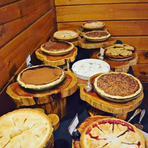 A variety of pies displayed on wooden stands, including chocolate, pecan, and fruit pies, against a rustic wooden backdrop.