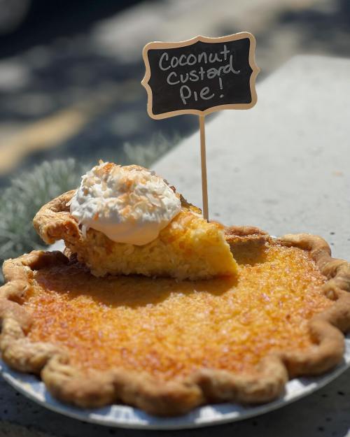 A coconut custard pie with a slice removed, topped with whipped cream and toasted coconut, displayed on a table.