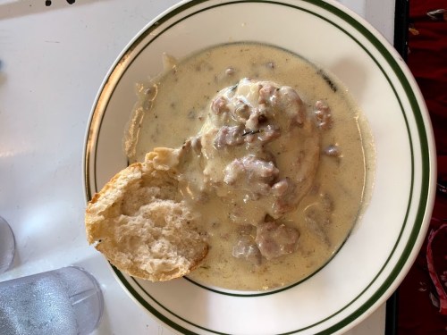 A plate of creamy sausage gravy over biscuits, with a piece of biscuit on the side.