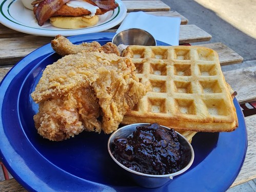 A plate featuring crispy fried chicken, a waffle, and a small cup of berry jam on a wooden table.