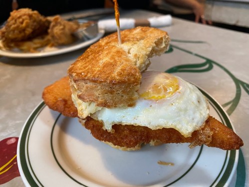 A fried chicken sandwich with a fried egg on a biscuit, served on a plate with a side of fried chicken in the background.