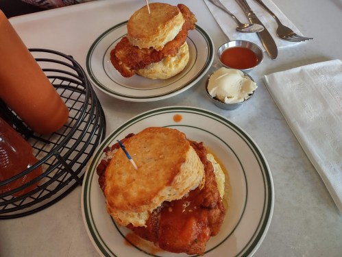 Two chicken biscuit sandwiches with sauce, served on plates with a side of butter and a bottle of hot sauce.