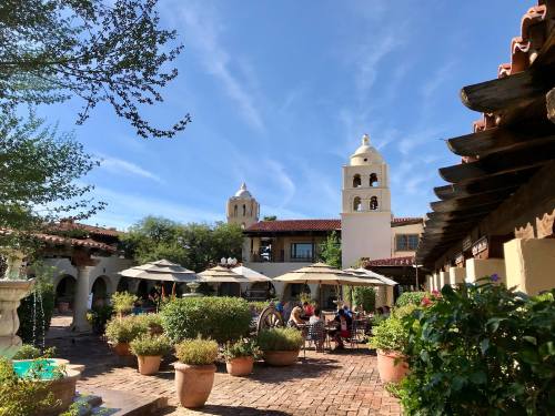 A sunny courtyard with potted plants, umbrellas, and people dining, framed by buildings with bell towers and blue sky.