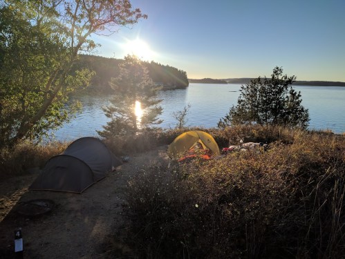 Two tents set up by a tranquil lakeside at sunset, surrounded by trees and tall grass.